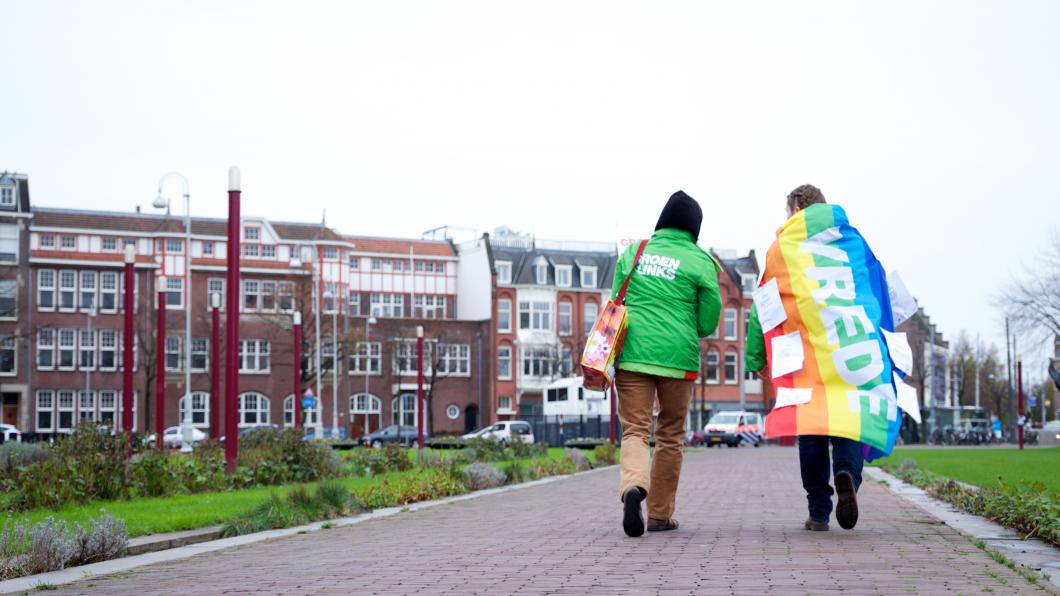 Twee vrijwilligers lopen over straat, een heeft de regenboogvlag met het woord 'vrede' over de schouders geslagen.